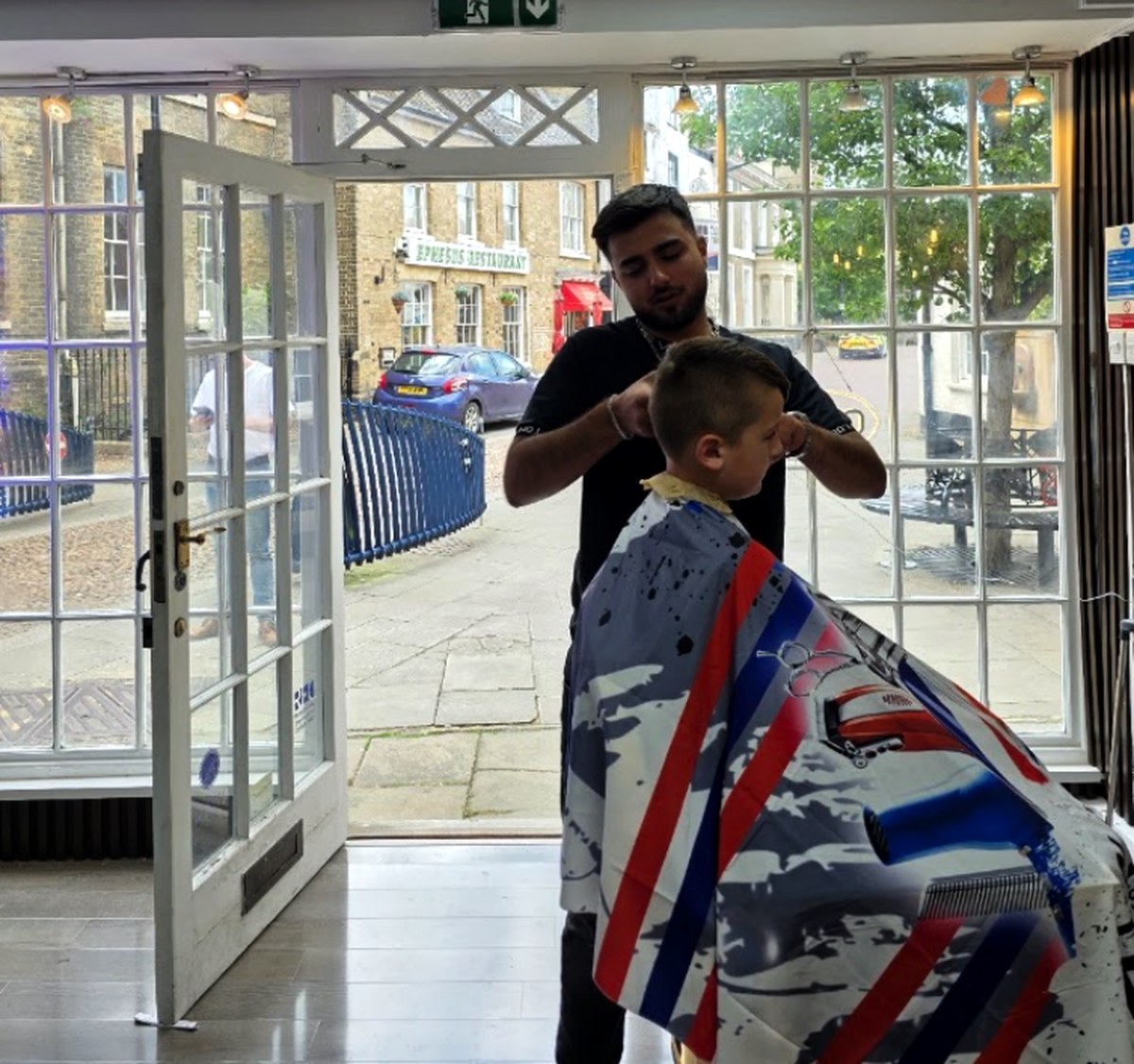 Barber at Huntingdon Barbers cutting a young client's hair by the shop window on Market Hill